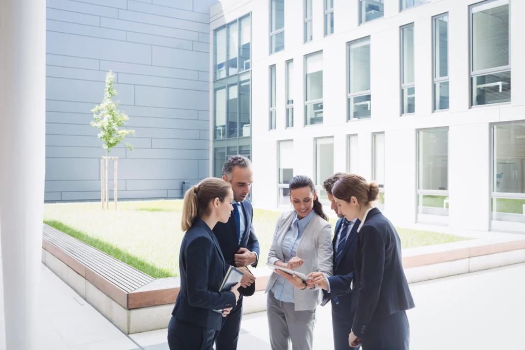 Group of five professionals in business attire gathered outdoors, looking at a tablet together in a modern office courtyard.