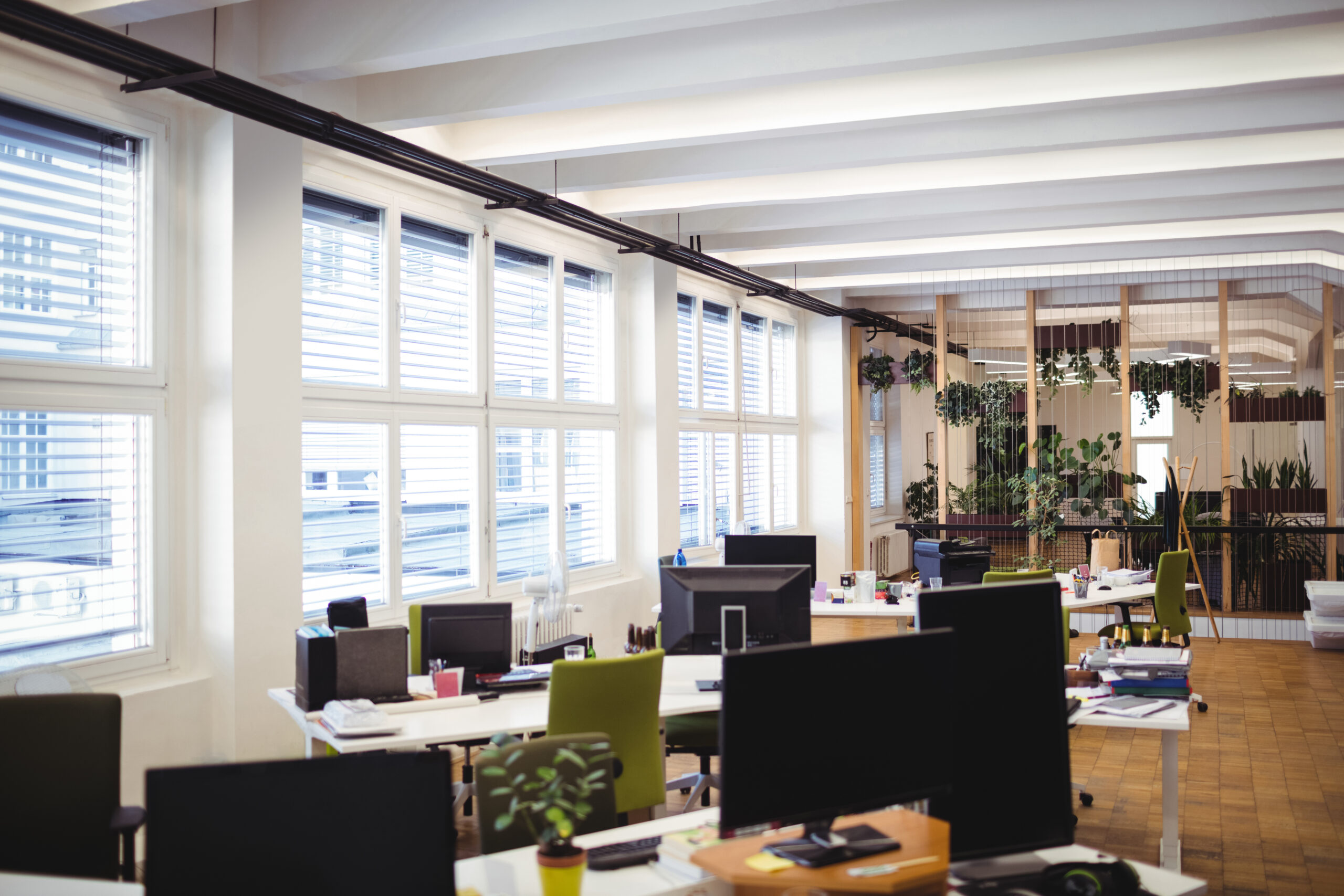 Empty office workplace with table chair and computer - The Gibson Company Open-plan office with multiple desks, computers, and potted plants by large windows with blinds.
